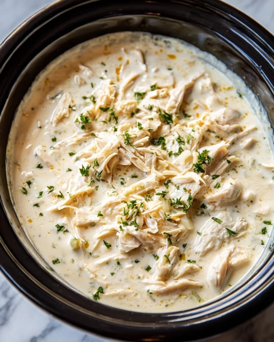 A black slow cooker filled with creamy chicken soup. The soup has a thick, white cream base and is topped with shredded white chicken pieces spread on the surface. Small bits of green fresh herbs are sprinkled evenly on top. The texture looks smooth and rich with soft chicken layers resting gently in the creamy sauce. The background is a white marbled surface. Photo taken with an iphone --ar 4:5 --v 7