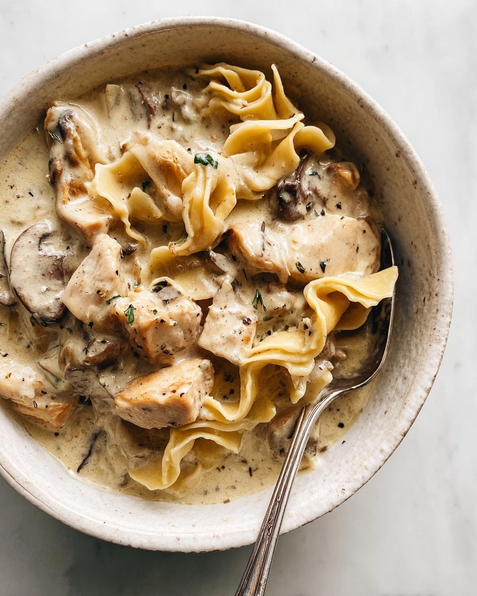 A rough-textured white bowl holds creamy mushroom pasta with a thick, pale beige sauce. The dish contains several layers: wide, ruffled sheets of pasta with a soft yellow color form the base, mixed with chunky pieces of light brown chicken and slices of dark brown mushrooms. The sauce coats everything evenly, showing small black herb bits and flecks of seasoning scattered throughout. A metal spoon rests inside the bowl, partially submerged in the rich sauce. The bowl sits on a white marbled surface. photo taken with an iphone --ar 4:5 --v 7