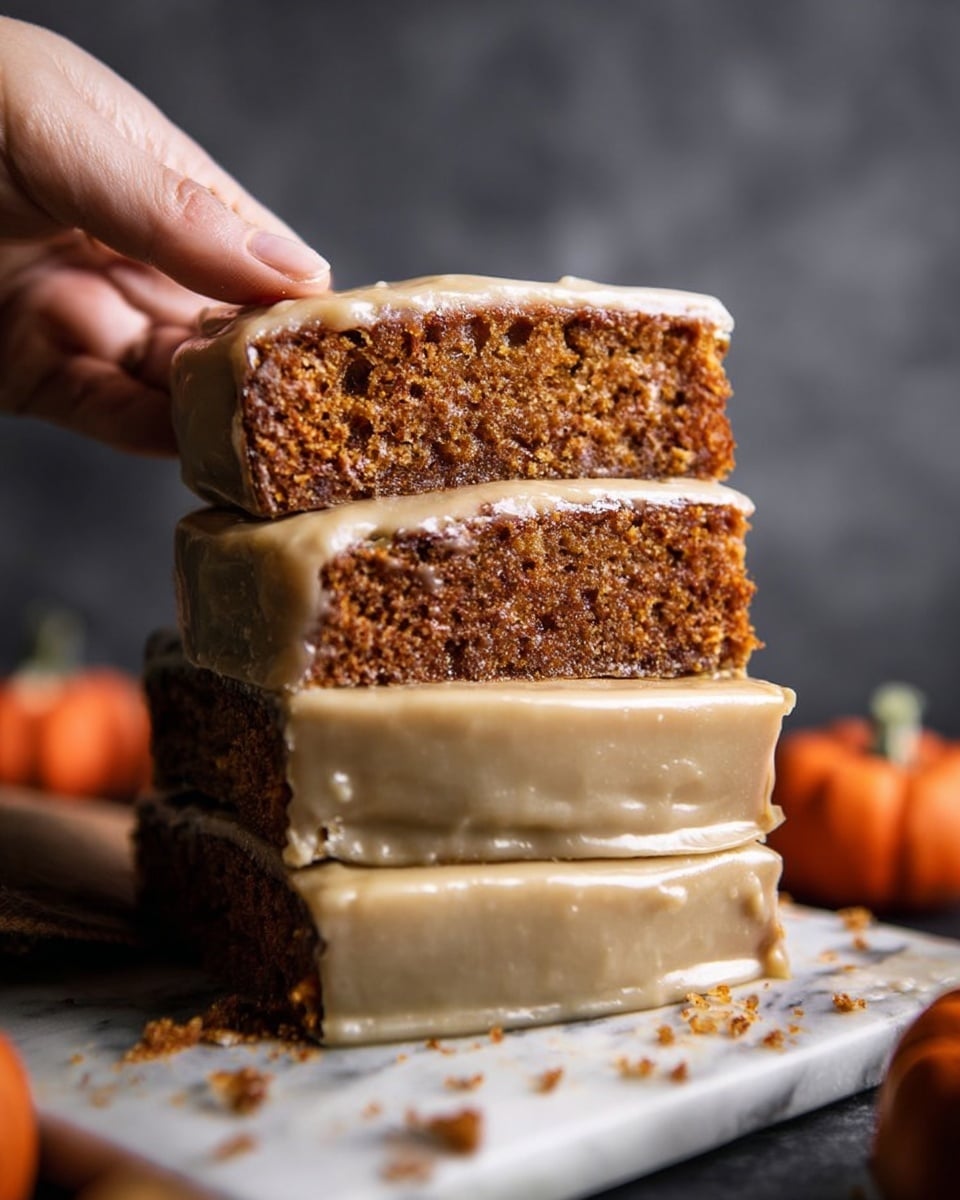 A thick stack of four slices of moist brown cake with smooth, glossy beige icing fully covering their sides is shown on a white marbled surface. Each cake slice layer is rich brown with a slightly crumbly texture at the top visible beneath the icing. A woman's hand is gently lifting the top slice, showing the thickness and soft texture of the cake. The background is blurred dark gray, with small pumpkins and scattered crumbs providing an autumn feel around the cake base. photo taken with an iphone --ar 4:5 --v 7