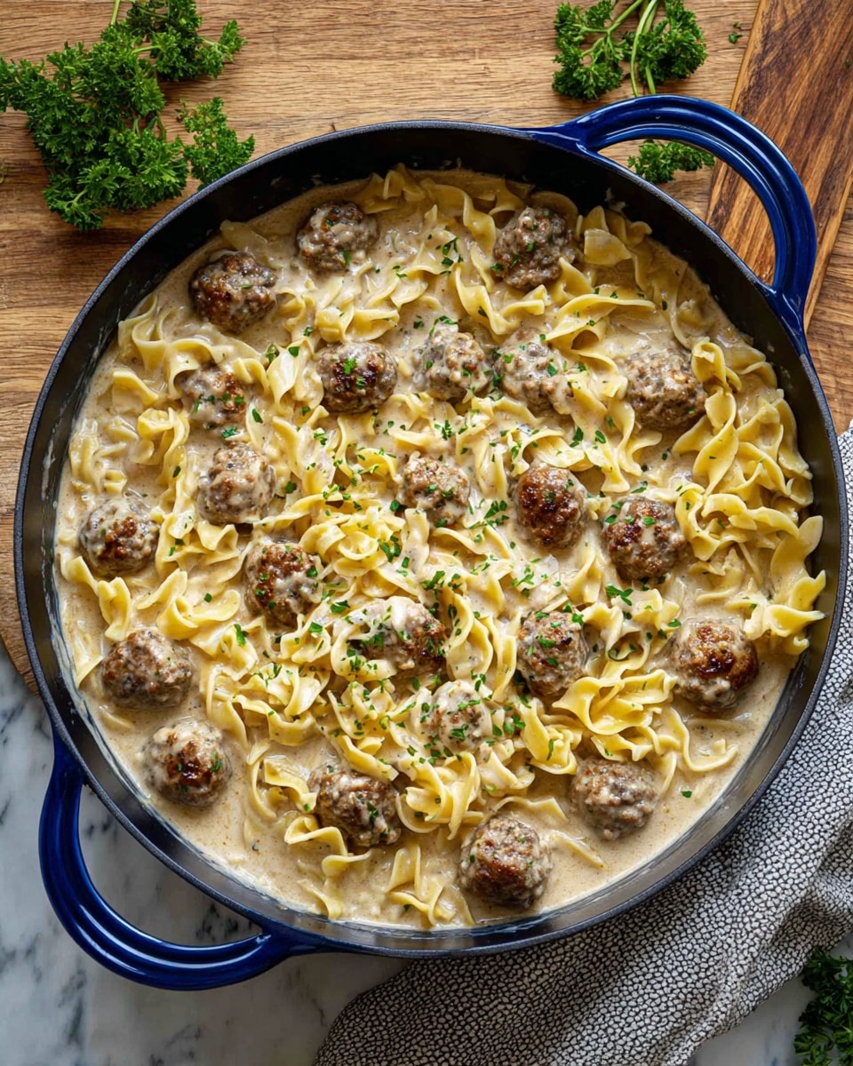 A large blue cast iron pan filled with a creamy beige sauce that covers the bottom layer with tender yellow egg noodles scattered throughout. On top, there are many small browned meatballs spread evenly across the pan, some partially covered in sauce. The dish is garnished with small bits of green herbs sprinkled over the top, adding a touch of color. The pan is placed on a wooden surface with a light gray textured cloth to the side and a few sprigs of green parsley around, all set on a white marbled background. Photo taken with an iphone --ar 4:5 --v 7