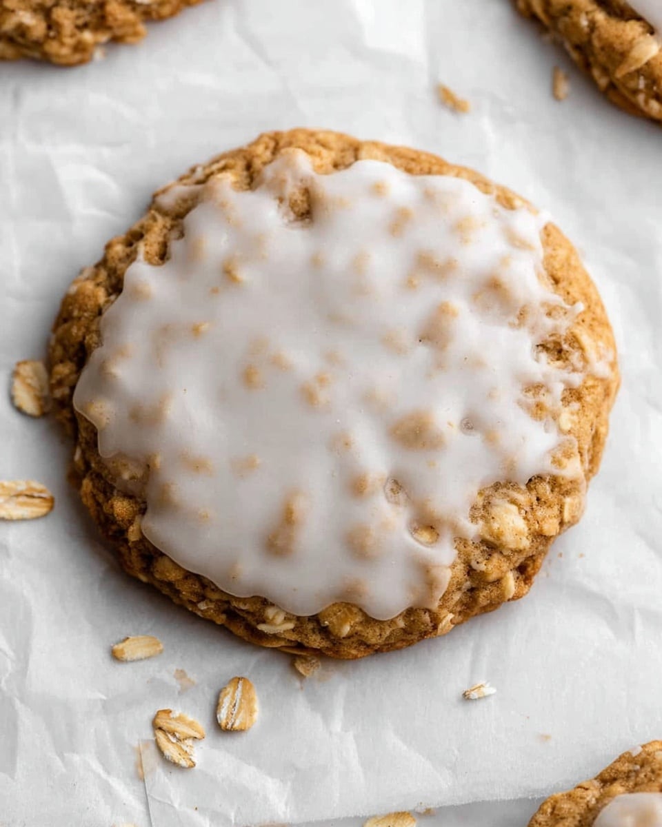 A single iced oatmeal cookie sits on a crinkled sheet of white parchment paper placed on a white marbled surface. The cookie has one main layer, a golden brown base with visible oat pieces giving it a textured look. On top, there is a smooth white icing that spreads unevenly, partly covering the oats underneath and blending softly into the edges. Some scattered oat flakes are near the cookie on the parchment paper, adding a natural element to the scene. photo taken with an iphone --ar 4:5 --v 7