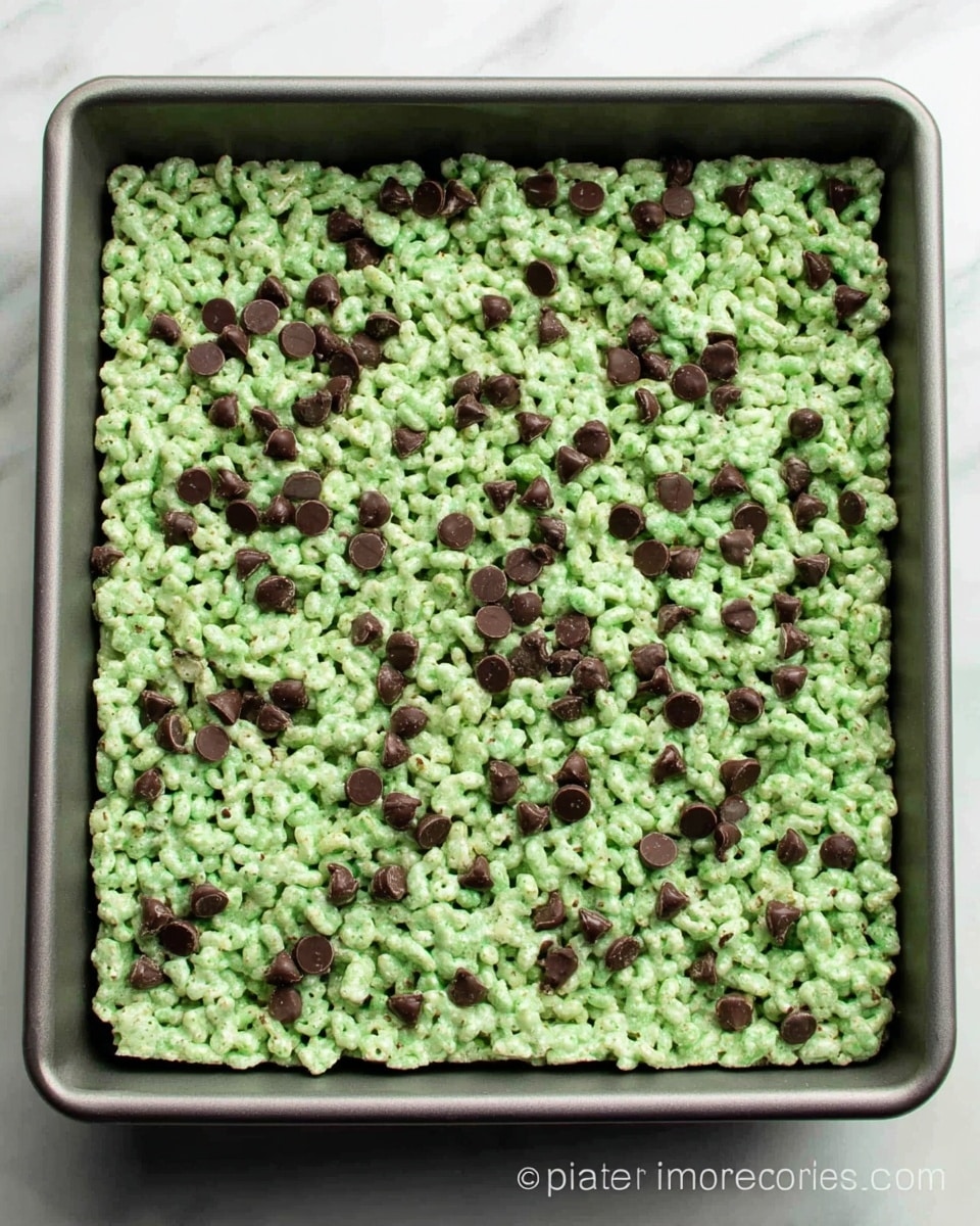 A square pan filled with a single layer of light green crispy cereal treats mixed evenly with small dark brown chocolate chips scattered all over. The cereal treats have a slightly sticky and shiny texture holding the cereal pieces together, forming a rough, bumpy surface. The pan is gray and placed on a white marbled surface. photo taken with an iphone --ar 4:5 --v 7