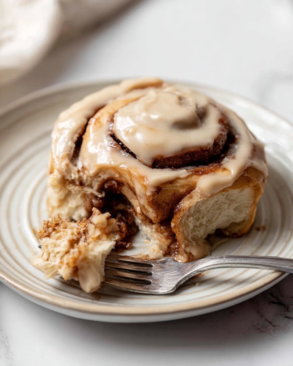 A close-up image of a cinnamon roll with soft, light brown dough twisted into a spiral shape, showing a darker cinnamon sugar filling between the layers. The roll is topped with a smooth, light tan cream cheese frosting that covers the top and drips slightly down the sides. One piece is cut and resting on a silver fork in front of the roll. Both the roll and the fork sit on a white plate with a subtle lined pattern, placed on a white marbled surface. photo taken with an iphone --ar 4:5 --v 7