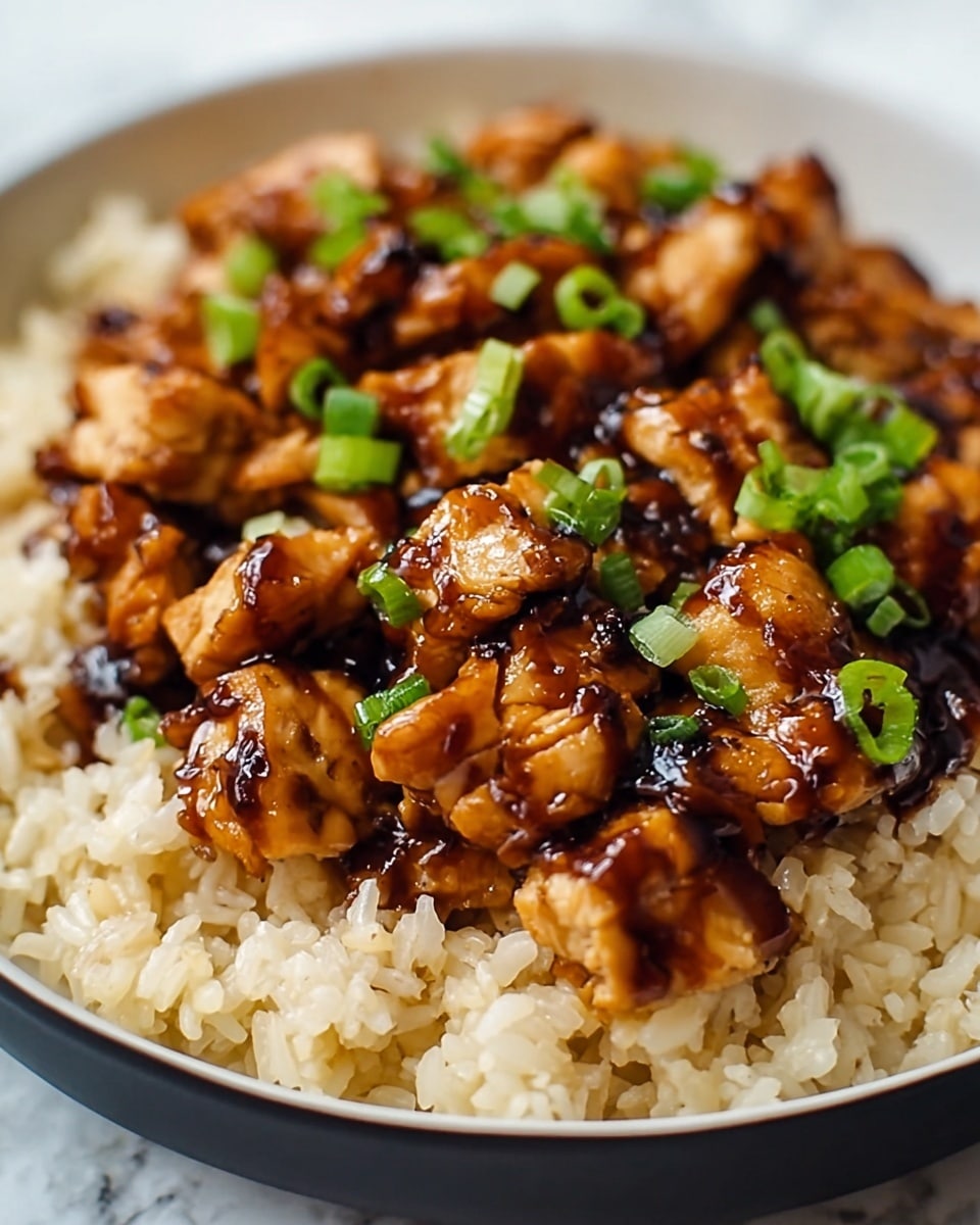 A close-up view of a dish in a white bowl showing two main layers: the bottom layer is light brown cooked rice with a soft and fluffy texture, and the top layer consists of small pieces of grilled chicken coated in a shiny dark brown glaze with slight char marks. Scattered across the chicken are small, bright green chopped spring onions adding color contrast. The bowl sits on a white marbled surface. photo taken with an iphone --ar 4:5 --v 7