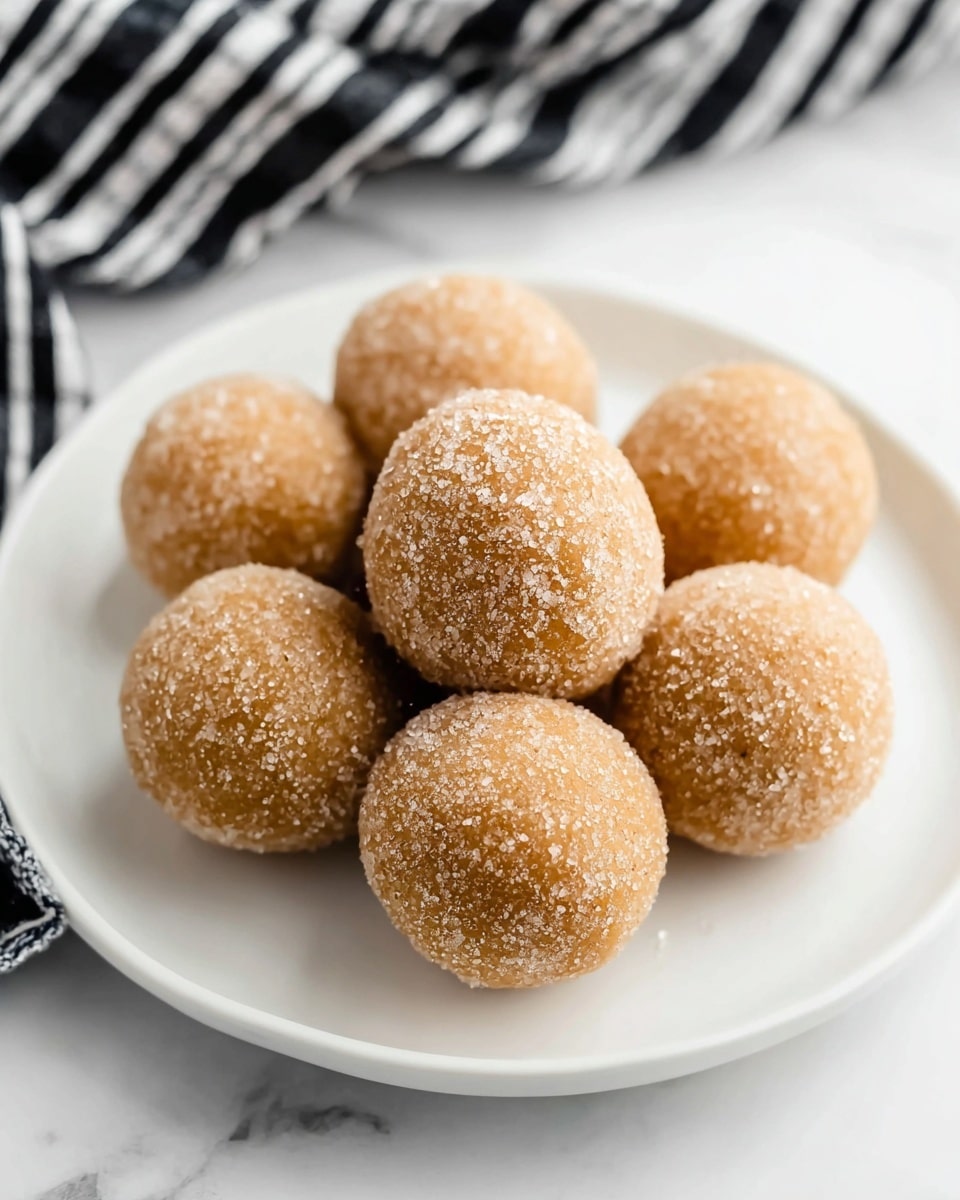 Seven round balls covered with granulated sugar are neatly arranged on a white plate. Each ball has a light brown color and a slightly rough texture from the sugar coating. The balls are placed close together in a circular pattern with one ball in the center and the others surrounding it. The plate sits on a white marbled surface, and a black and white striped cloth is blurred in the background. photo taken with an iphone --ar 4:5 --v 7