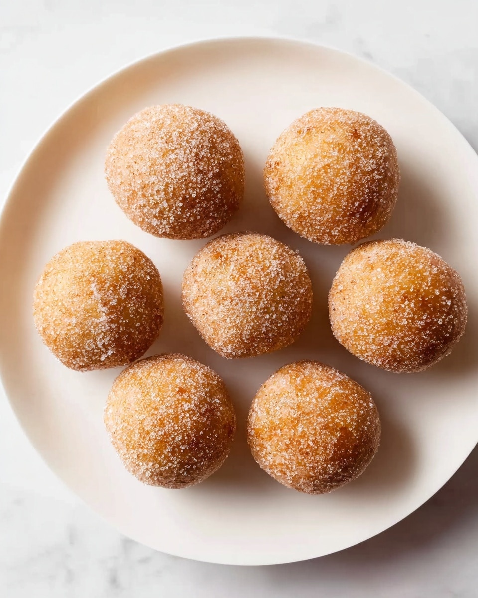 Eight round sugar-coated doughnut holes are arranged in a circle on a plain white plate. Each doughnut ball is light golden brown with a rough sugar grain texture all over its surface, giving a slightly sparkly look. The plate sits on a white marbled surface, and soft natural lighting highlights the sugary texture and the smooth, evenly browned dough underneath. The shadows underneath each doughnut are faint, adding depth but keeping focus on the sweet balls. photo taken with an iphone --ar 4:5 --v 7