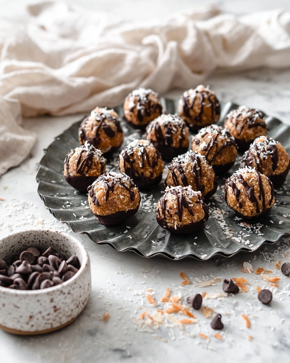 Round energy balls are arranged on a dark grey fluted metal tray on a white marbled texture. Each ball has a base layer dipped in dark chocolate, a middle layer of light brown nutty texture with small bits, topped with a drizzle of dark chocolate and sprinkled with white toasted coconut flakes. Scattered chocolate chips and toasted coconut flakes surround the tray. In the foreground left, a white speckled bowl holds extra dark chocolate chips. A light beige fabric is softly draped in the background. Photo taken with an iphone --ar 4:5 --v 7