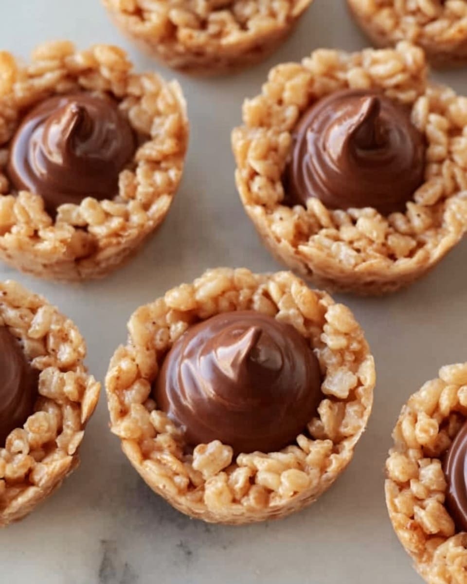 The image shows several small round treats made from a crispy light brown puffed rice base shaped into cups, each filled with a smooth, glossy layer of rich milk chocolate in the center. The treats are closely arranged on a white marbled surface. The chocolate filling has a soft texture with slight swirls on top, contrasting with the crunchy look of the puffed rice cups around it. photo taken with an iphone --ar 4:5 --v 7