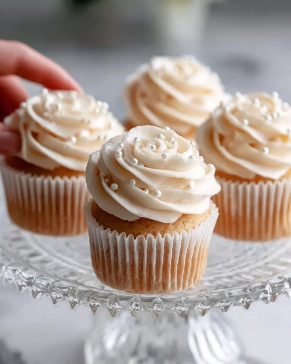 The image shows four cupcakes on a clear glass cake stand, each topped with a swirl of light cream-colored frosting shaped like a rose with small pearl-like sprinkles scattered on top. The cupcakes have a light brown base with white paper liners around them. The background is a white marbled texture, and a woman’s hand is gently touching one of the cupcakes on the stand. photo taken with an iphone --ar 4:5 --v 7