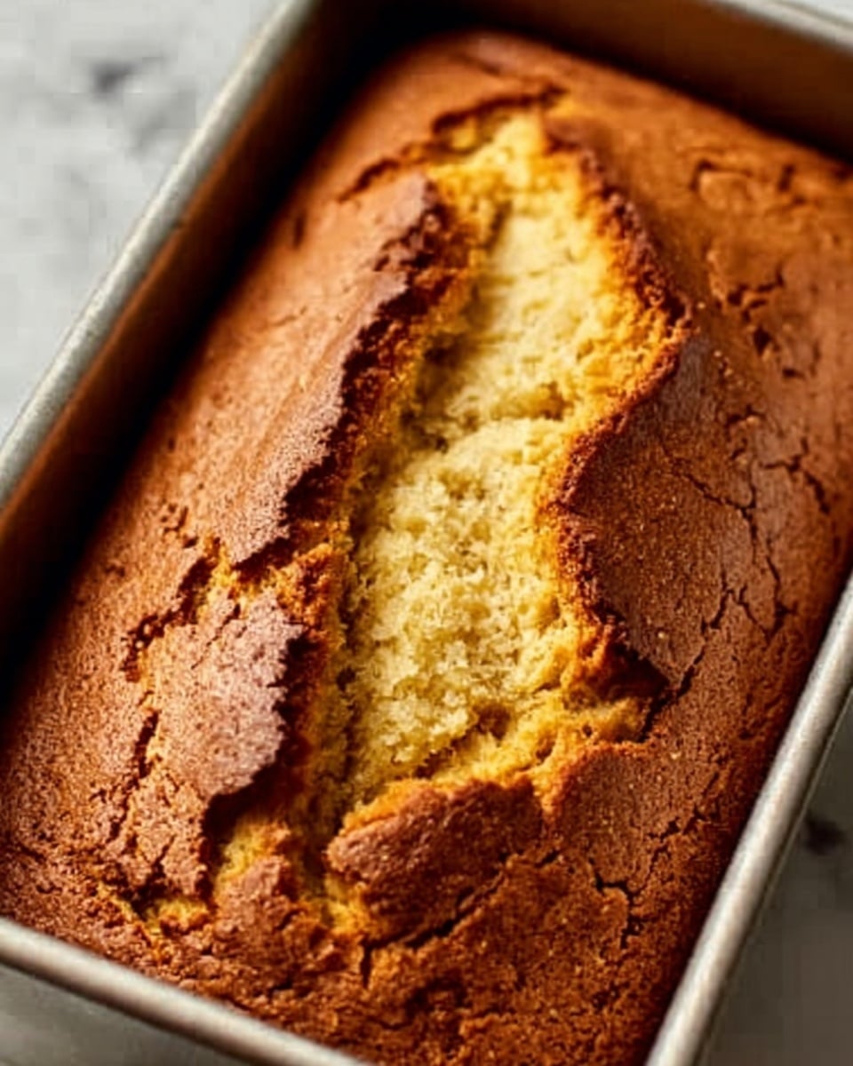 A close-up image of a golden brown loaf cake with a cracked top, sitting inside a metal baking pan. The cake has a slightly rough, crumbly texture with small cracks showing its soft inside. The background is a white marbled texture. Photo taken with an iphone --ar 4:5 --v 7