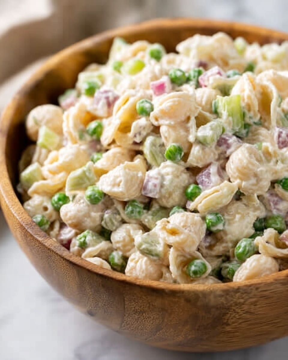 A close-up image of a wooden bowl filled with creamy pasta salad. The salad has three visible layers: small spiral and shell pasta in light yellow coated with white creamy dressing, scattered green peas adding bright spots of color, and small pieces of red onion and celery providing purple and pale green bits throughout. The bowl sits on a white marbled surface, slightly blurred in the background, drawing attention to the colorful pasta salad in the foreground. photo taken with an iphone --ar 4:5 --v 7