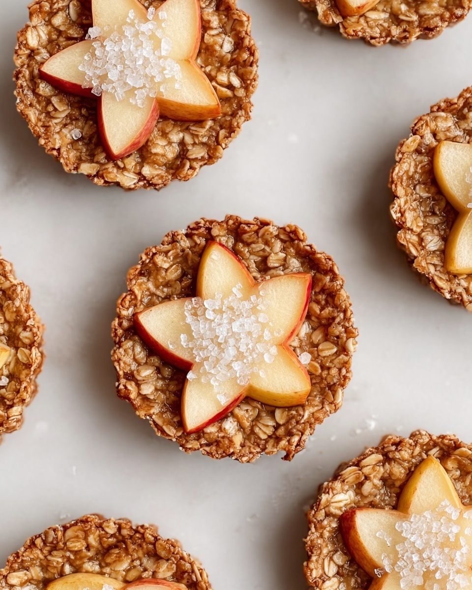 The image shows small round oat tarts arranged on a white marbled surface. Each tart has a textured, golden-brown oat crust that looks soft and chewy. On top, there are four thin apple slices arranged in a star shape with the skin on the edges adding a touch of red and yellow color. The center of each tart is sprinkled with coarse white sugar crystals, creating a sparkling effect against the warm tones of the oats and apples. The tarts are evenly spaced, and the photo is taken with an iphone --ar 4:5 --v 7