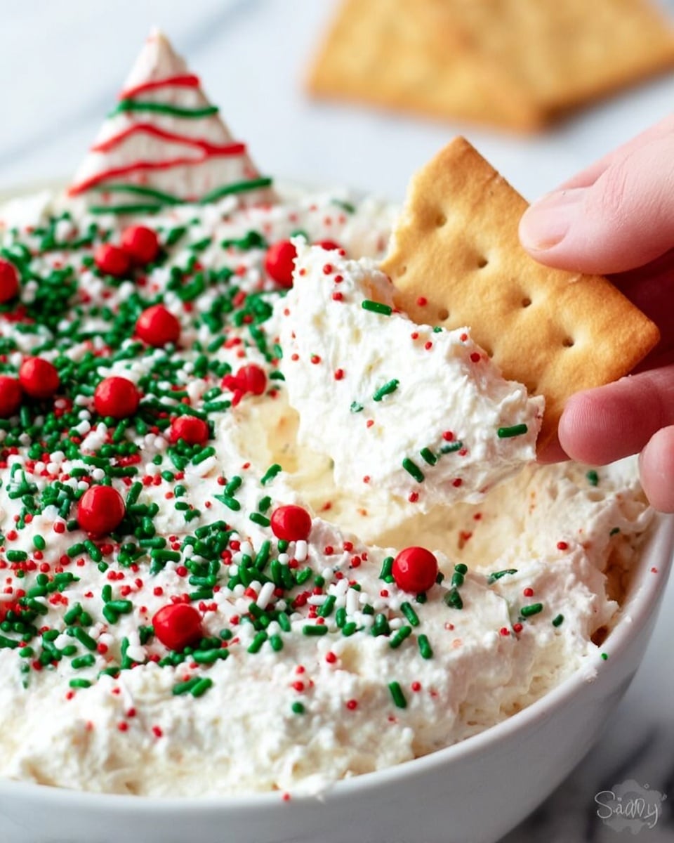 A close-up image shows a white bowl filled with creamy white dip that looks fluffy and soft. The dip is decorated with two types of sprinkles: small round red balls and longer green sprinkles, spread evenly on top. In the dip, a piece of white triangular chip with red and green lines is stuck upright near the back edge of the bowl. A woman's hand holds a light brown rectangular cracker dipped into the creamy mixture, with the dip clinging to the cracker along with some sprinkles. The bowl sits on a white marbled surface. photo taken with an iphone --ar 4:5 --v 7