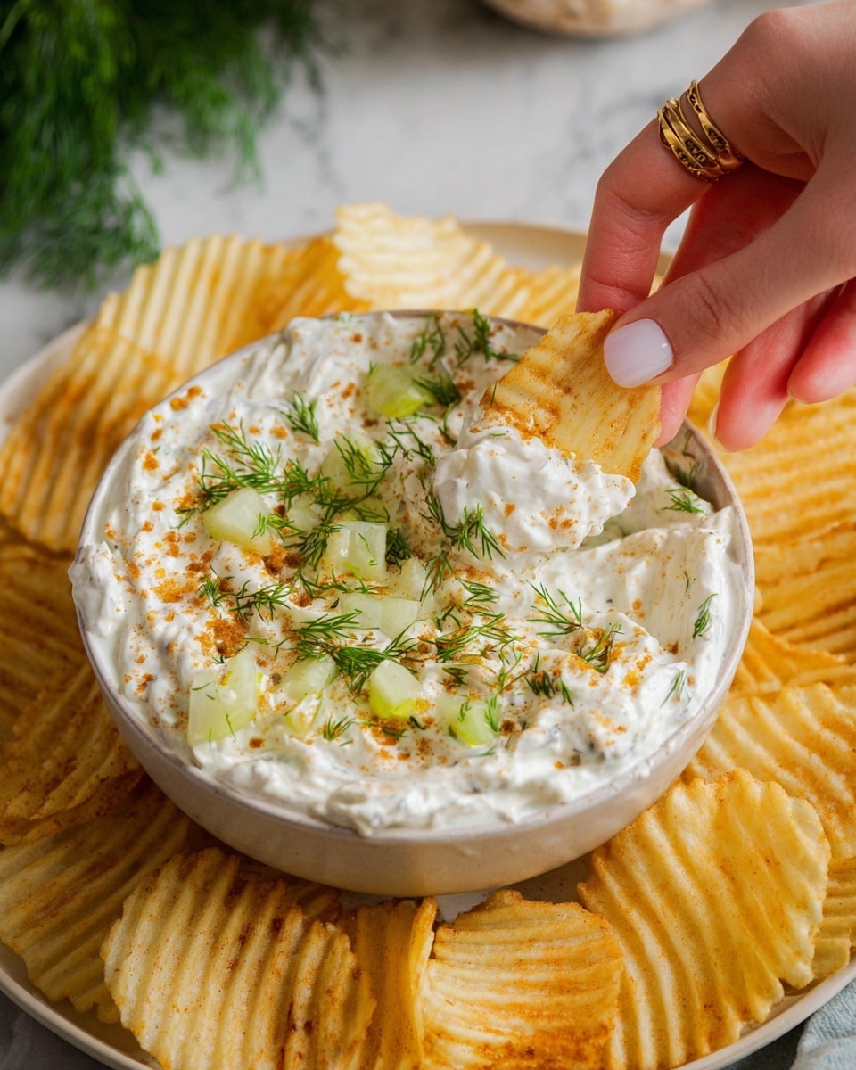 The image shows a close-up of a bowl of creamy white dip with a slightly rough texture. The dip is topped with small green pickle pieces, bright green dill sprigs, and a light orange powder sprinkled lightly over the surface. A woman's hand is holding a ridged potato chip dipped in the creamy mixture, with some dip visible on the chip's edge. The white bowl sits on a white plate that holds many golden-yellow ridged potato chips all around it. The setting is on a white marbled surface with fresh green dill partially visible in the background. photo taken with an iphone --ar 4:5 --v 7