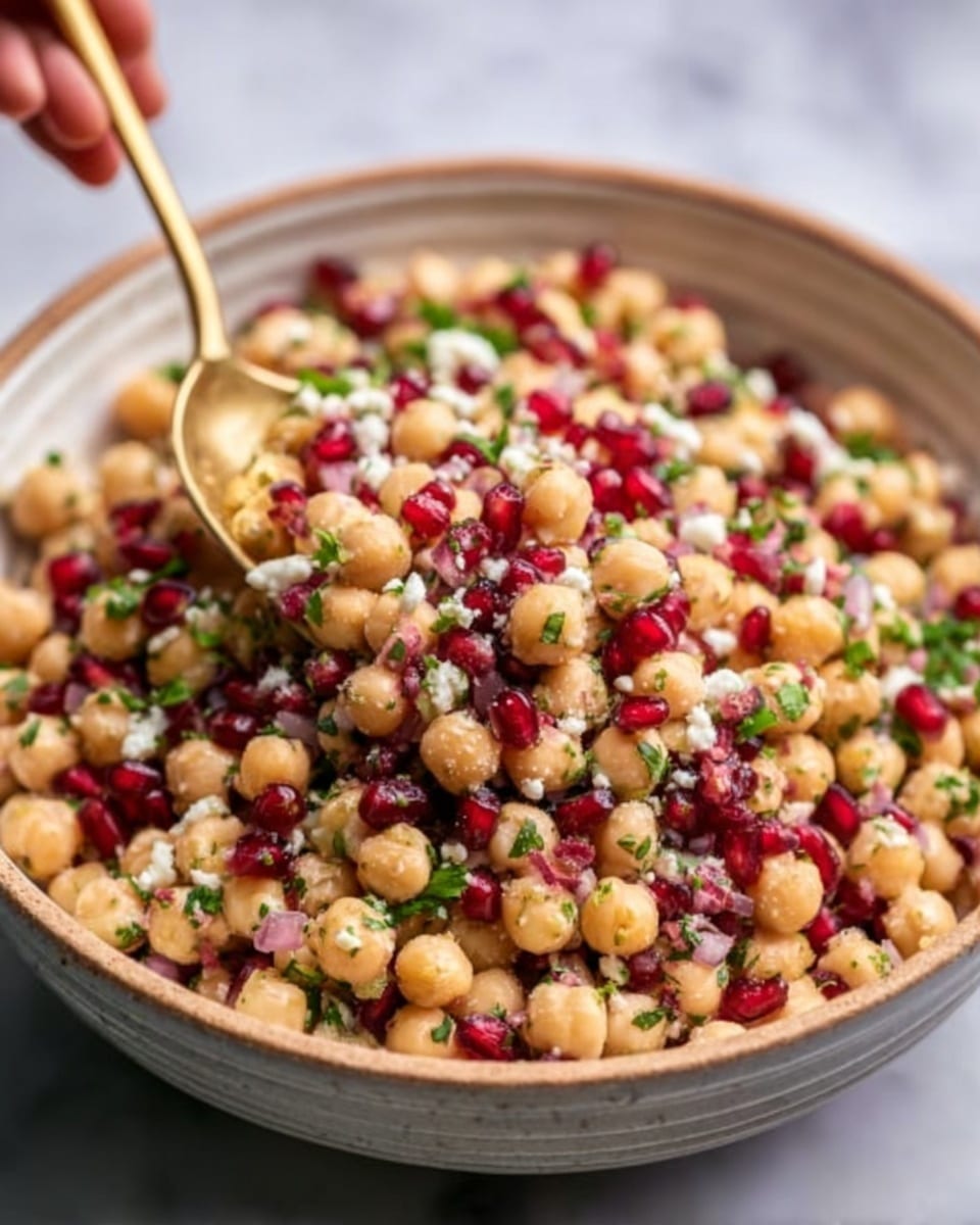 A white bowl filled with a colorful chickpea salad sits on a white marbled surface. The salad has three main layers: the base layer is light beige chickpeas, the middle layer includes bright red dried cranberries scattered evenly, and the top layer is small pieces of white cheese mixed with green herbs sprinkled throughout. The chickpeas look smooth and round, the cranberries have a shiny and slightly wrinkled texture, and the cheese appears soft with small crumbly chunks. The overall look is vibrant and fresh. photo taken with an iphone --ar 4:5 --v 7