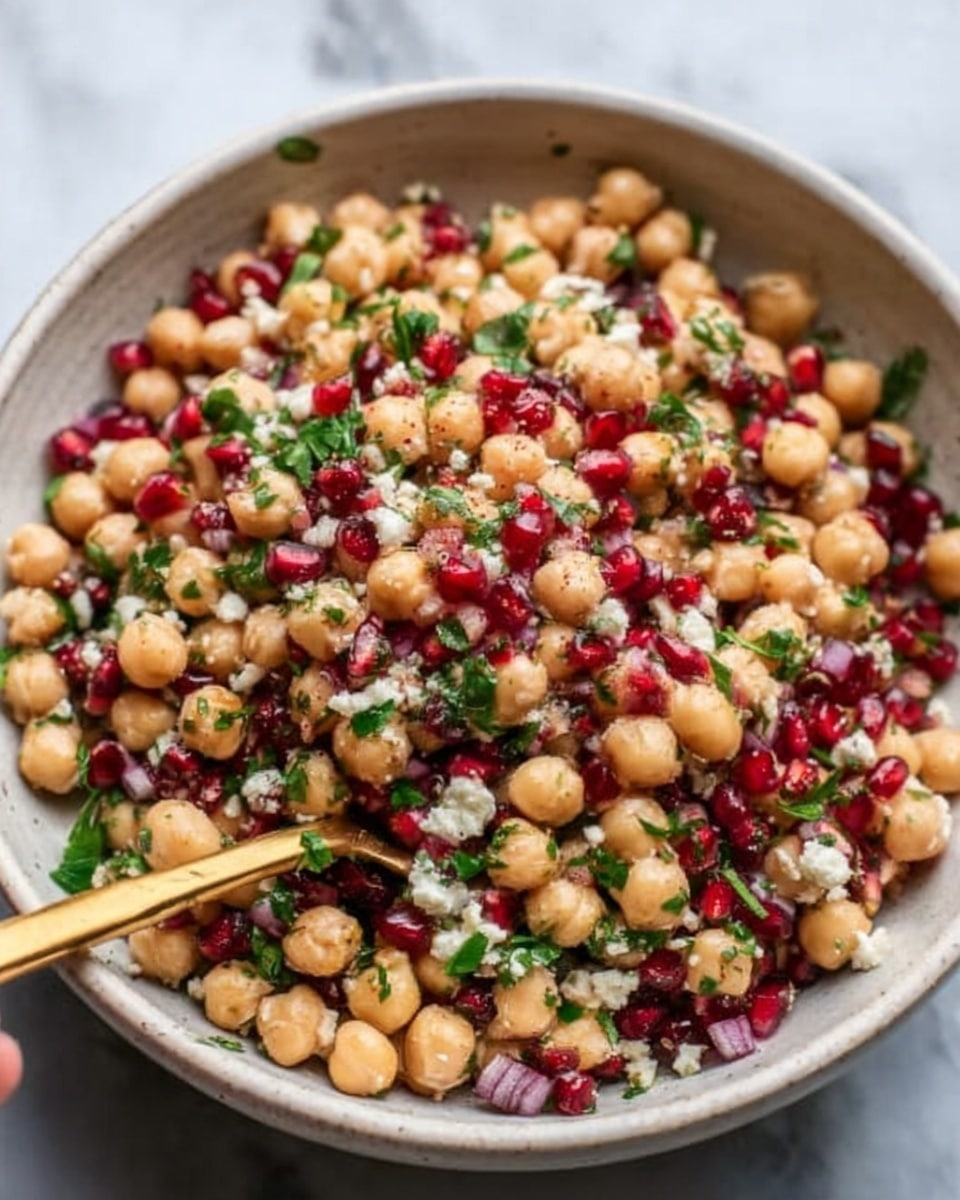 A bowl filled with a chickpea salad showing multiple layers of ingredients; the bottom layer is light beige chickpeas that look soft and round, mixed with small deep red pomegranate seeds scattered throughout, adding bright color contrast. Mixed within are small pieces of green herbs and tiny white crumbles of cheese on top. The bowl is white, sitting on a white marbled surface, and a woman's hand is holding a gold spoon digging into the salad. Photo taken with an iphone --ar 4:5 --v 7