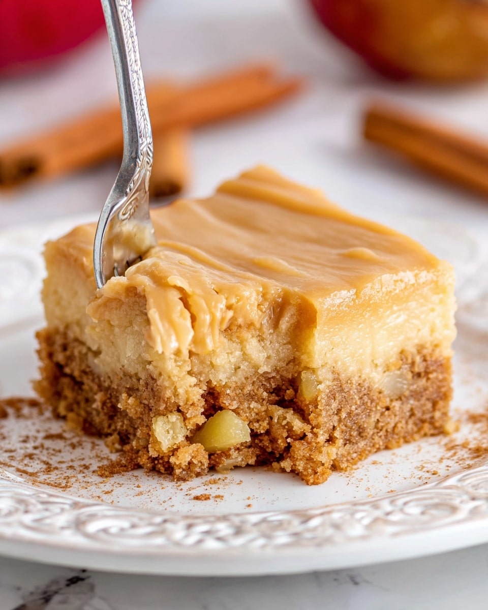The image shows a close-up of a square dessert with two visible layers on a white decorated plate placed on a white marbled surface. The bottom layer is thick, crumbly, and brown with bits of yellow chunks inside, giving it a textured, moist look. The top layer is smooth, glossy, and light caramel brown, forming a firm but soft finish. A silver fork is spearing a small piece off the front corner, highlighting the layers and texture. In the background, slightly blurred, there are warm brown cinnamon sticks and part of a red apple. Photo taken with an iphone --ar 4:5 --v 7