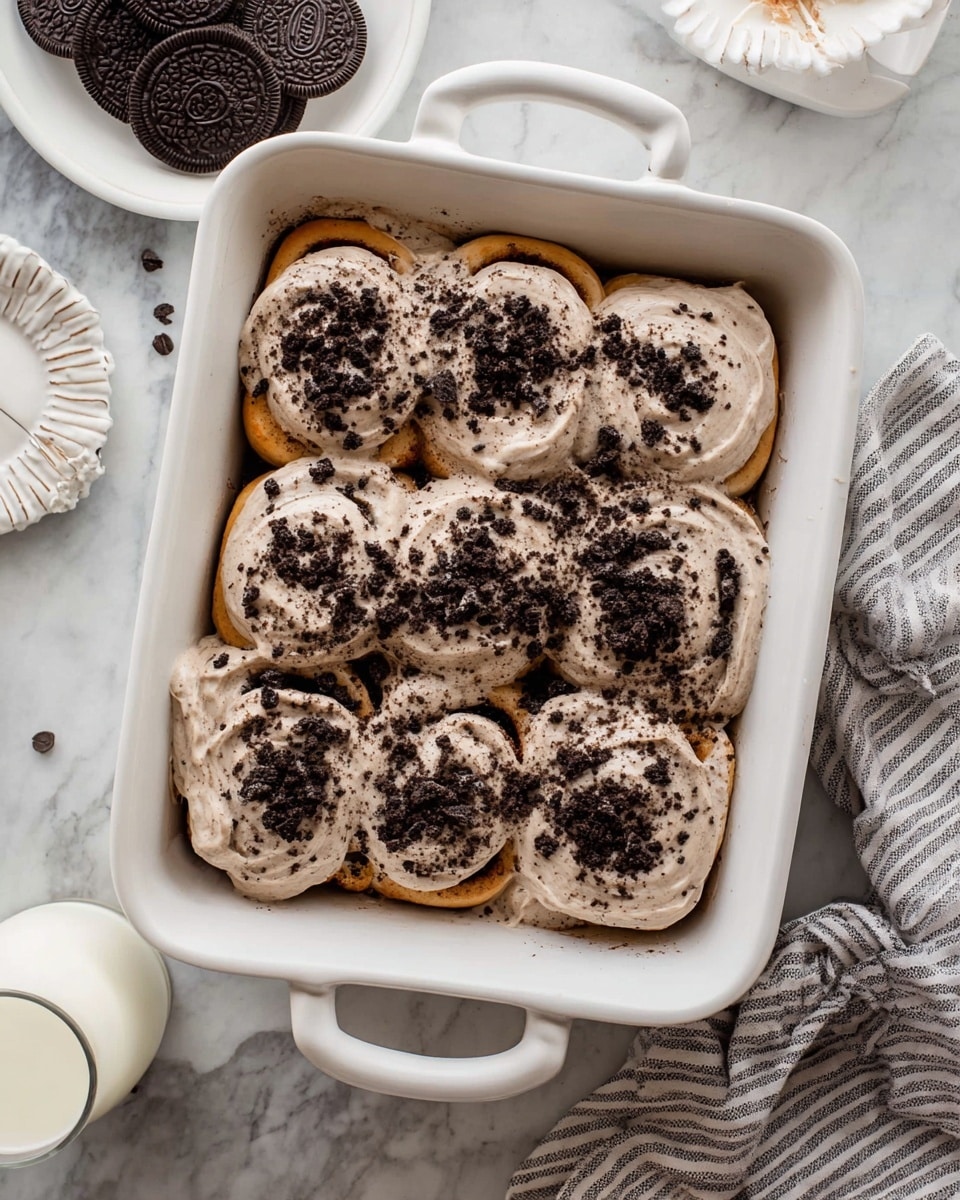 A white rectangular baking dish holds a batch of nine cinnamon rolls arranged in three rows of three. Each roll is covered with a thick layer of creamy white frosting mixed with crushed dark cookie crumbs, giving a speckled black and white top. The cinnamon rolls have a golden brown dough base partially visible underneath the frosting. The dish sits on a white marbled texture surface next to a small white bowl of dark cookie pieces, an empty white scalloped plate, two glasses of milk, and a gray and white striped cloth. Photo taken with an iphone --ar 4:5 --v 7