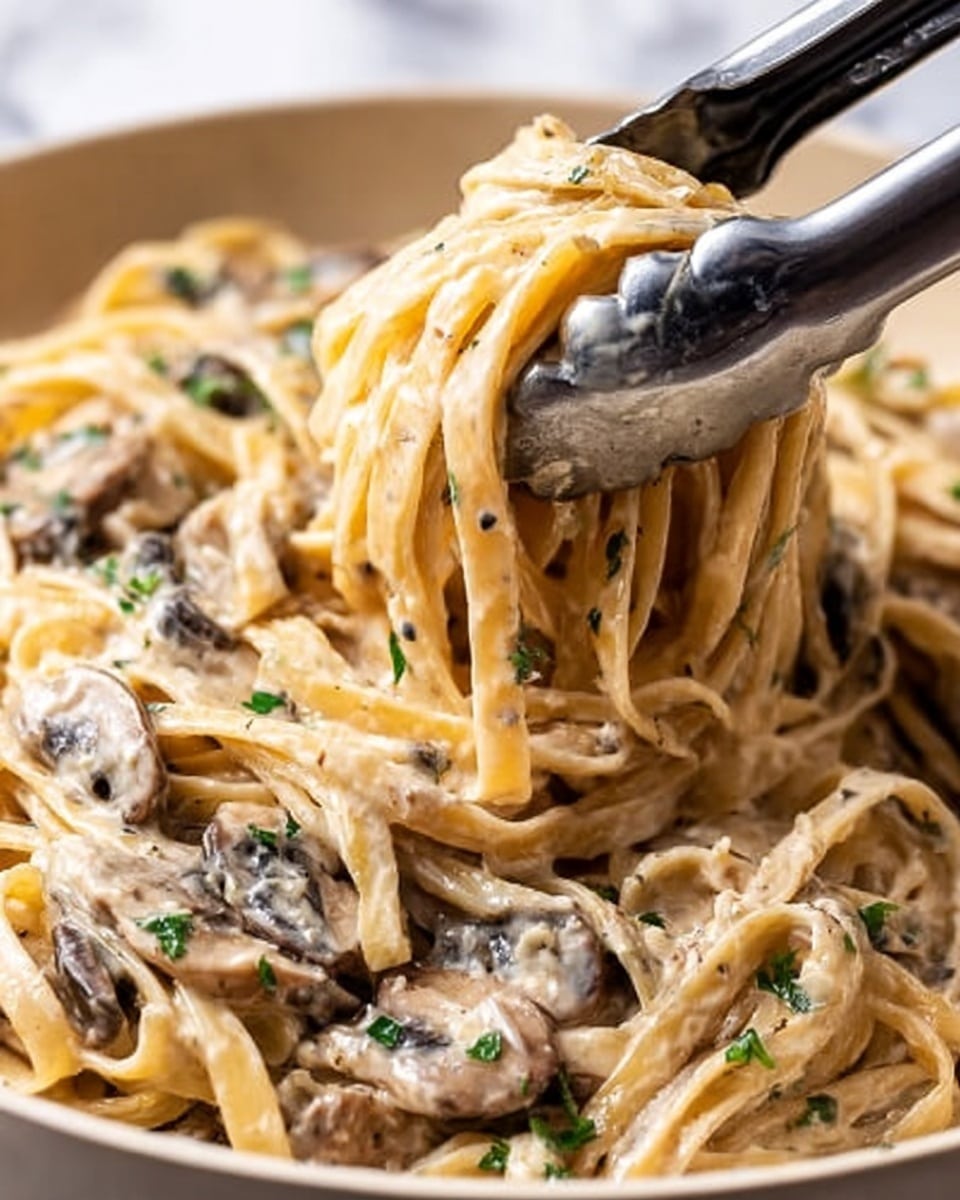 A close-up view of creamy fettuccine pasta mixed with sliced mushrooms, coated in a thick, light beige sauce. The noodles are soft and tangled together with small green parsley pieces scattered throughout. A black and silver spatula lifts a portion of the pasta, showing the smooth texture of the sauce and the tender mushrooms nestled among the noodles. The background is a white marbled texture. photo taken with an iphone --ar 4:5 --v 7