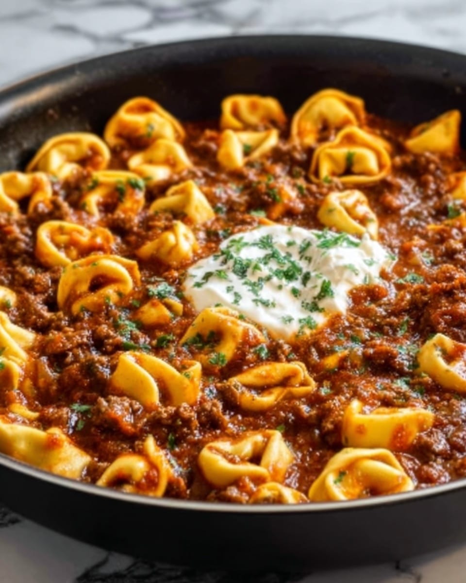 A close-up view of a large black pan filled with many small tortellini pasta pieces covered in thick, rich brown meat sauce. On top, there is a small dollop of white cream and a sprinkle of green herbs. The tortellini have a yellow, soft texture, and the sauce looks chunky and hearty, with bits of meat visible. The pan sits on a white marbled surface. Photo taken with an iphone --ar 4:5 --v 7