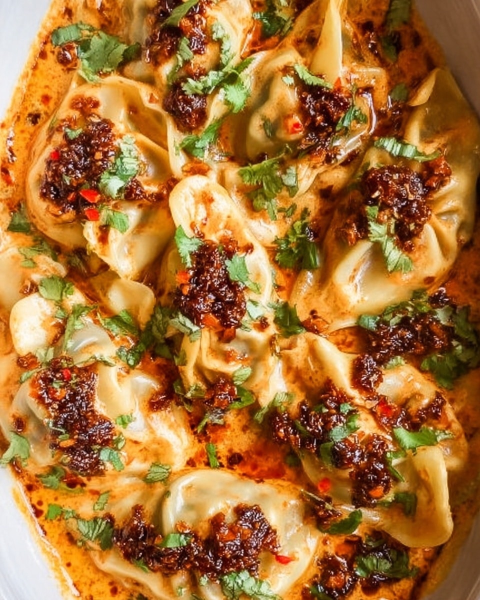 A white dish filled with soft pasta dumplings layered closely together, covered in a creamy orange sauce with oil spots that have a reddish tint. Each dumpling is topped with a chunky dark brown garlic and chili paste, garnished with bright green cilantro leaves scattered around. The sauce has a smooth texture with small bits of herbs mixed in. The overall look is warm and spicy, with the dumplings showing soft folds and some areas lightly browned. The dish rests on a white marbled surface. photo taken with an iphone --ar 4:5 --v 7