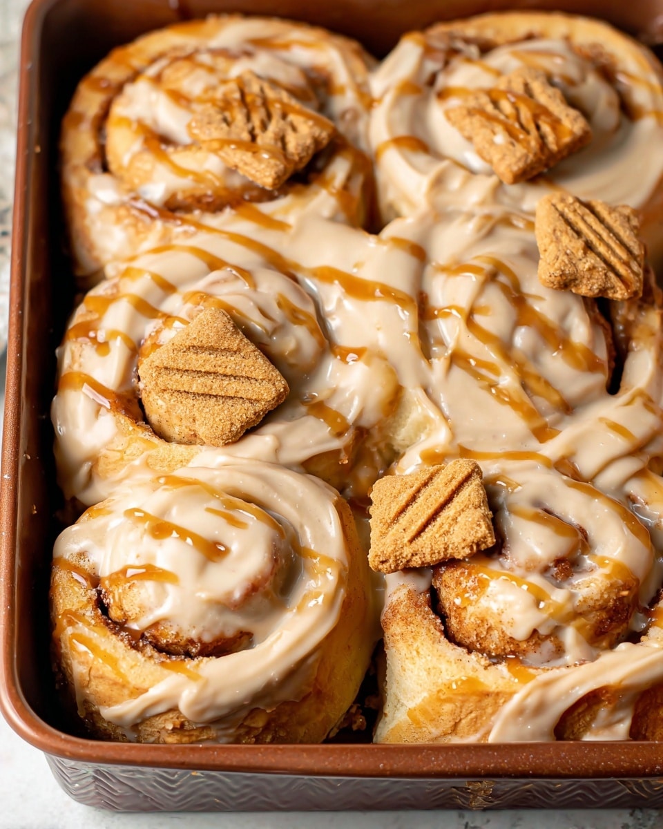 The image shows a close-up of six cinnamon rolls covered in a thick, creamy beige glaze with drizzles of caramel sauce on top. Each cinnamon roll has a slightly browned, soft, and swirled dough texture visible beneath the glaze. There are small rectangular brown cookies with an embossed design placed on three of the cinnamon rolls, adding contrast. The rolls are in a dark brown baking pan with a handle, and the background features a white marbled texture. photo taken with an iphone --ar 4:5 --v 7