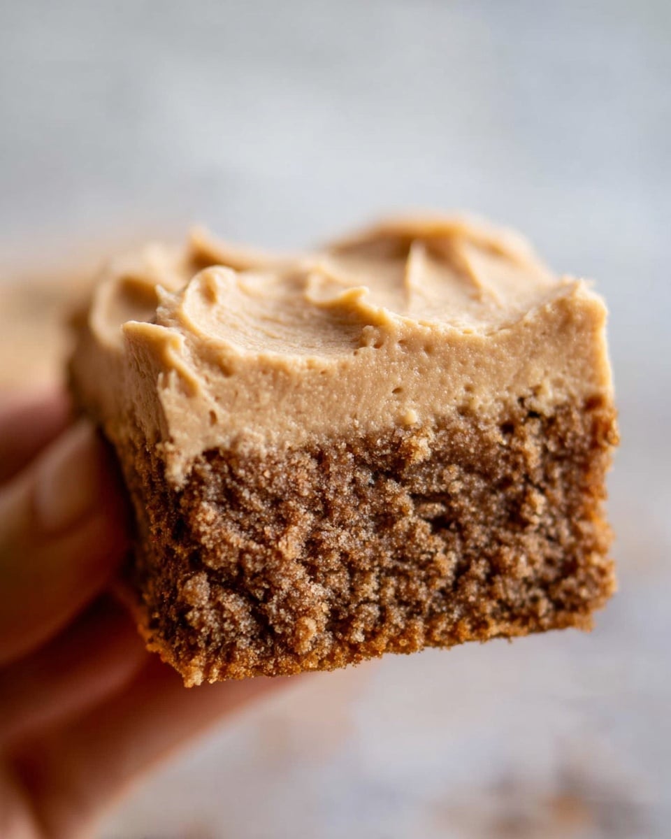 A close-up of a square piece of cake held by a woman's hand, showing two clear layers. The bottom layer is thick, moist, and crumbly with a brown color and small dark spots throughout. The top layer is a smooth, creamy frosting in a light tan color with a textured surface showing soft peaks and swirls. The background has a blurred white marbled texture. photo taken with an iphone --ar 4:5 --v 7