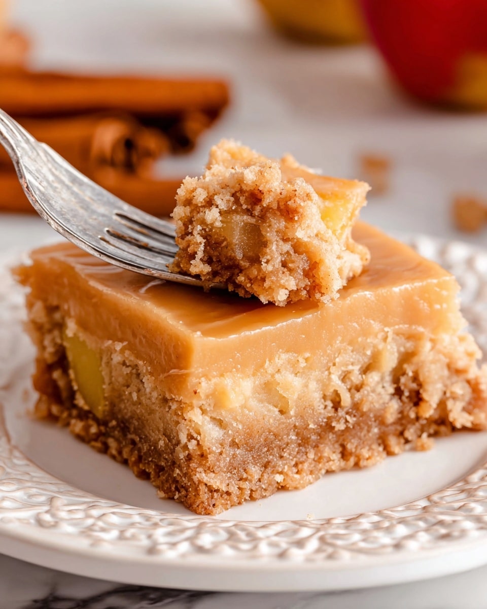 The image shows a square piece of dessert with two visible layers on a white plate with detailed embossed patterns. The bottom layer is crumbly and brown with small chunks of yellow fruit mixed in, giving it a rough texture. The top layer is smooth and light caramel-colored, covering the whole piece evenly. A silver fork is pressing into one corner, lifting part of the dessert to reveal its softness and moist crumbly texture. In the background, there are blurred elements in warm brown tones, suggesting cinnamon sticks and some red fruit. The whole setting is on a white marbled surface. photo taken with an iphone --ar 4:5 --v 7