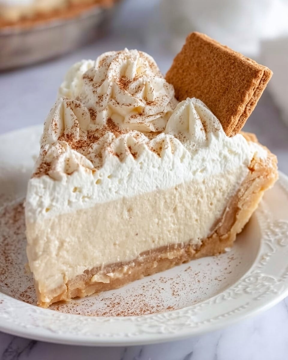 A slice of pie is shown on a white plate with a delicate textured edge, placed on a white marbled surface. The pie has three main layers: a golden light-brown flaky crust at the bottom and sides, a thick creamy beige filling in the middle, and a generous layer of white whipped cream on top. The whipped cream is swirled and sprinkled lightly with cinnamon powder. A square piece of graham cracker is inserted vertically into the whipped cream near the back edge of the slice. Photo taken with an iphone --ar 4:5 --v 7