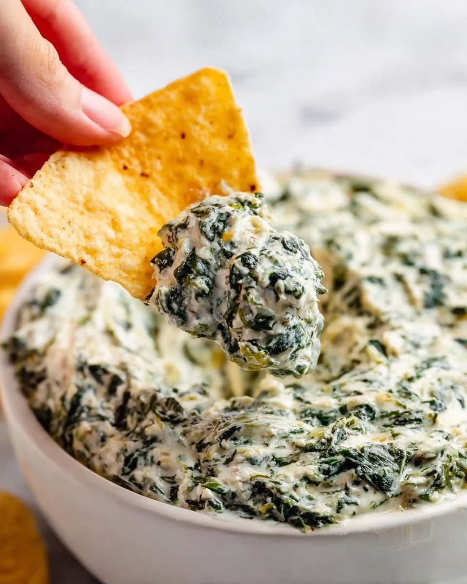 A close-up image shows a white bowl filled with creamy spinach dip that has a textured mix of dark green spinach leaves and white creamy sauce. In the foreground, a woman's hand holds a triangular, light yellow tortilla chip topped with a generous scoop of the same dip, highlighting the dip's thick and chunky texture with visible spinach pieces. The background and surface are a white marbled texture, creating a clean and bright look. photo taken with an iphone --ar 4:5 --v 7
