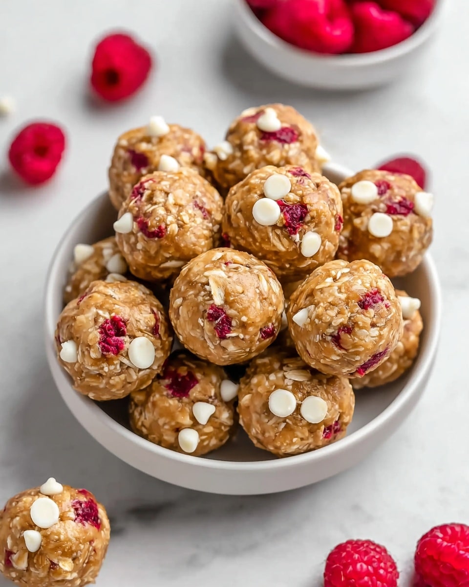 A light beige bowl filled with round energy balls made of oats and a brown dough mix, each ball showing white oats and small bright red dried berry pieces embedded in the surface, some balls topped with small white chocolate chips. Around the bowl, there are separate small white bowls holding fresh red raspberries, all set on a white marbled texture surface. Photo taken with an iphone --ar 4:5 --v 7