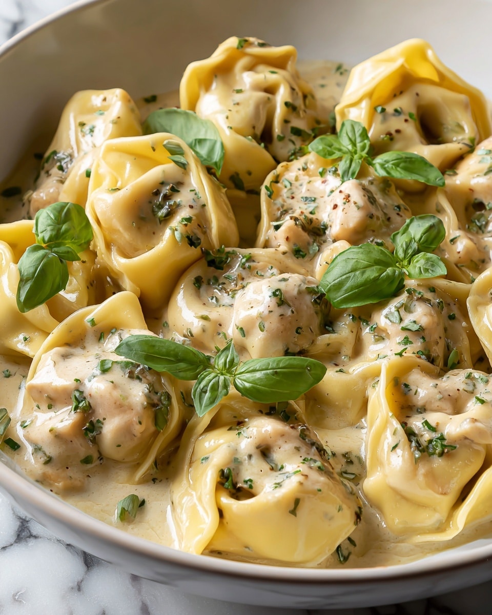The dish shows a close-up of several tortellini pasta pieces filled with a creamy chicken mixture, each pasta having a smooth light yellow color. They sit in a rich, light beige cream sauce that looks thick and slightly glossy. Small green basil leaves are scattered on top, adding bright color and freshness, while tiny bits of green herbs are sprinkled over both the sauce and pasta. The tortellini are arranged closely together, filling the whole white bowl, placed on a white marbled surface. photo taken with an iphone --ar 4:5 --v 7