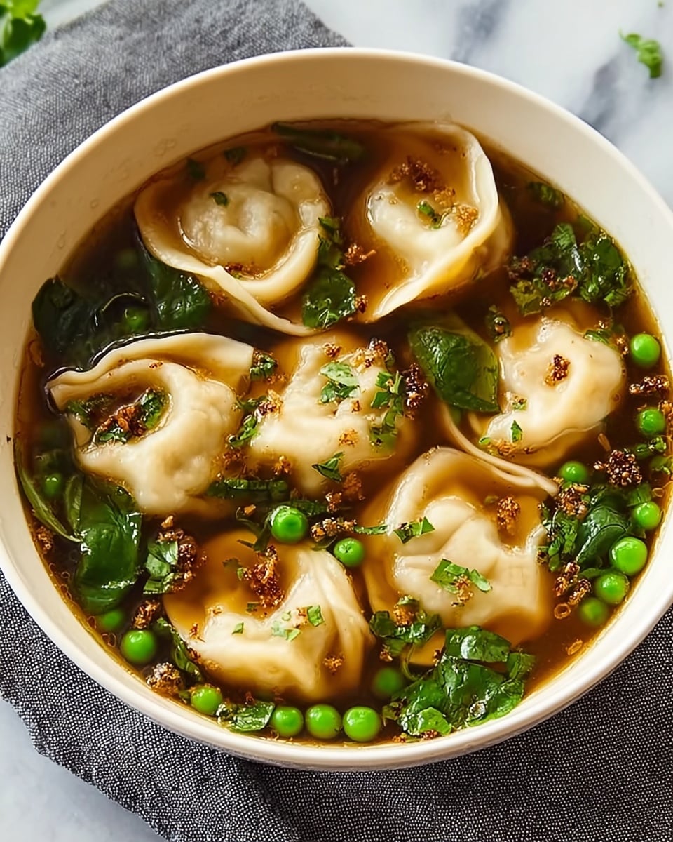 A white bowl with a thin black rim is filled with a clear brown broth. Inside the broth, there are multiple light beige dumplings with soft, folded edges floating on top. Scattered around the dumplings are small bright green peas and dark green leafy pieces. Tiny bits of light brown fried garlic or shallots and some black pepper flakes are sprinkled throughout. The bowl sits on a grey and white patterned cloth, with a blurred green leaf in the background on a white marbled texture. photo taken with an iphone --ar 4:5 --v 7