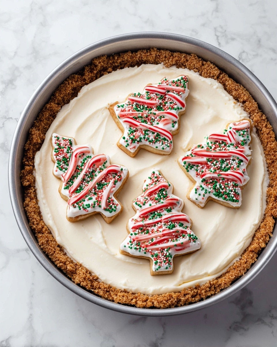 A round dessert with three visible layers in a gray springform pan on a white marbled surface; the bottom layer is a crumbly, light brown graham cracker crust pressed up the sides; the middle layer is a thick, smooth, creamy white filling covering the crust completely; the top layer has five tree-shaped cookies with white icing, red wavy lines, and small green sprinkles, arranged in a circle with a small gap in the middle, placed on the creamy layer. photo taken with an iphone --ar 4:5 --v 7