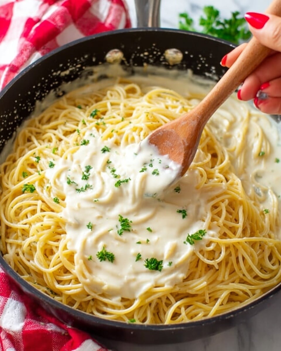 A close-up of a black frying pan filled with yellow cooked spaghetti topped with a creamy white sauce, sprinkled with small green parsley leaves. A woman's hand with red nail polish is stirring the pasta with a wooden spoon, lifting some sauce on the left side of the pan. The background shows a white marbled surface with some green herbs and a red checkered cloth blurred out. Photo taken with an iphone --ar 4:5 --v 7