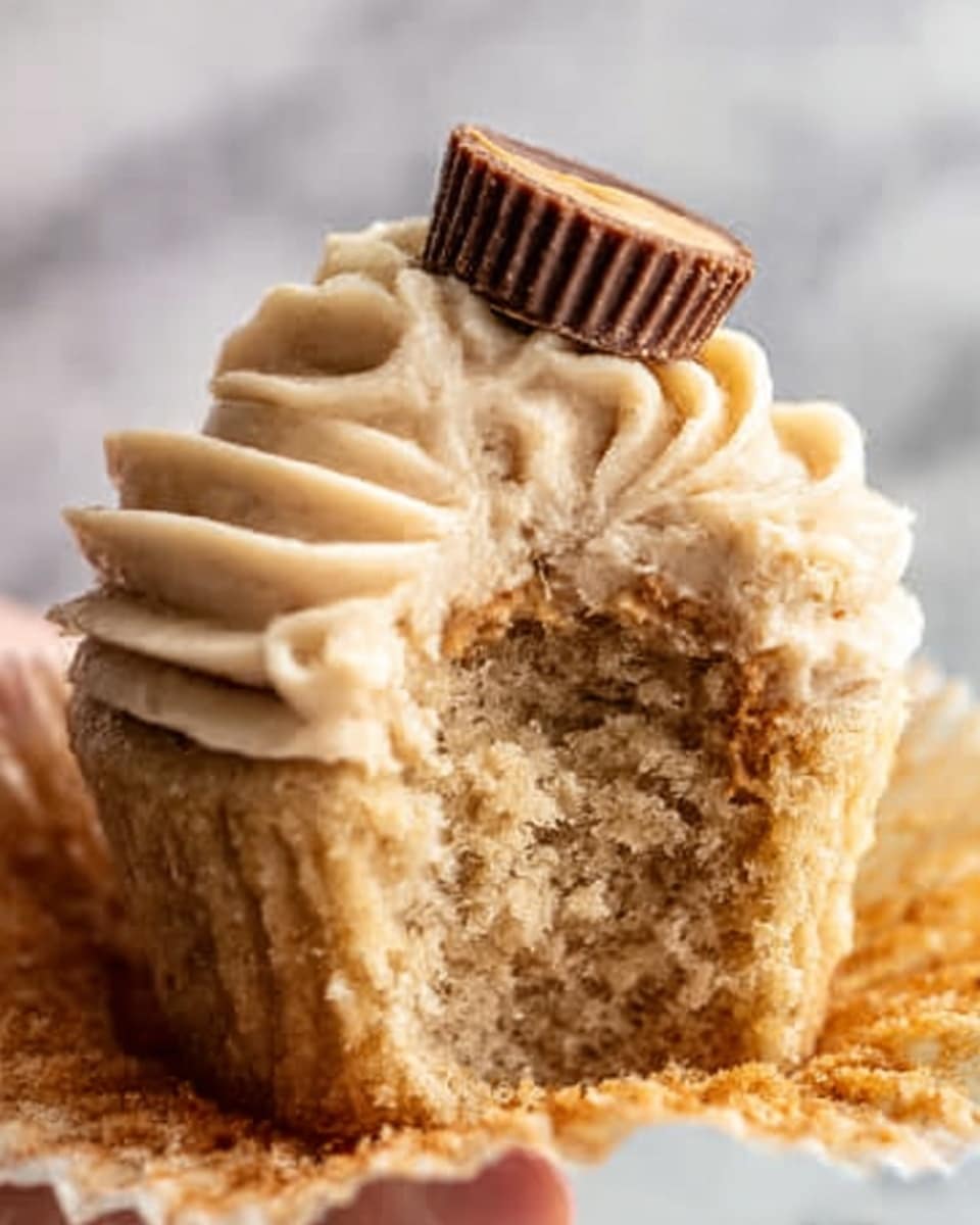 A close-up of a cupcake with one big bite taken out of it, showing a soft, fluffy light brown crumb inside. The cupcake is topped with a thick layer of smooth, pale beige frosting swirled neatly on top, and a small peanut butter cup nestled in the center of the frosting. The cupcake sits on a white plate with a white marbled surface in the background. Photo taken with an iphone --ar 4:5 --v 7