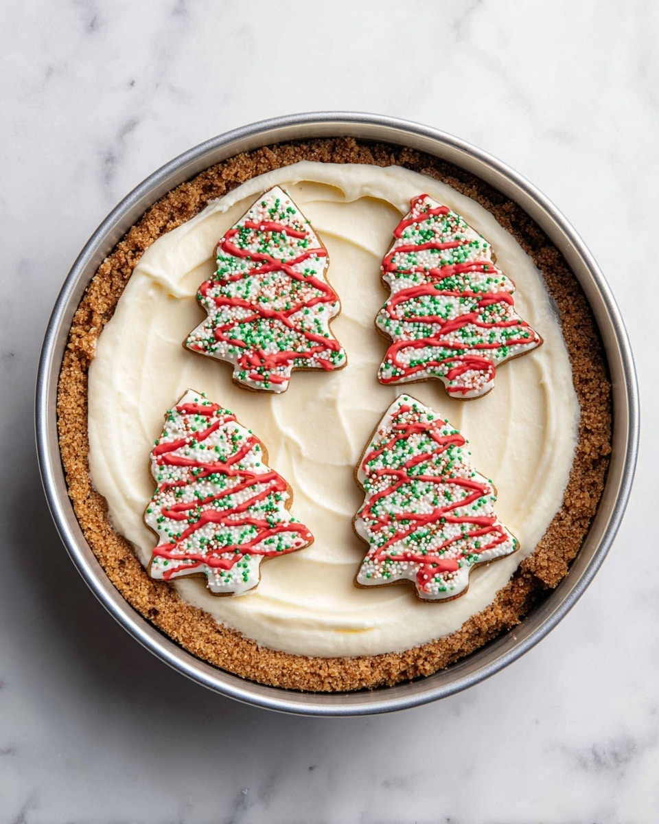 A round dessert with three visible layers in a metal pan: the bottom layer is a crumbly brown crust pressed against the sides, the middle layer is a smooth, creamy white filling spread evenly, and the top layer consists of five tree-shaped cookies with white icing, red wavy lines, and green sprinkles, placed in a circular pattern on the filling. The background surface is white with a marbled texture. Photo taken with an iphone --ar 4:5 --v 7