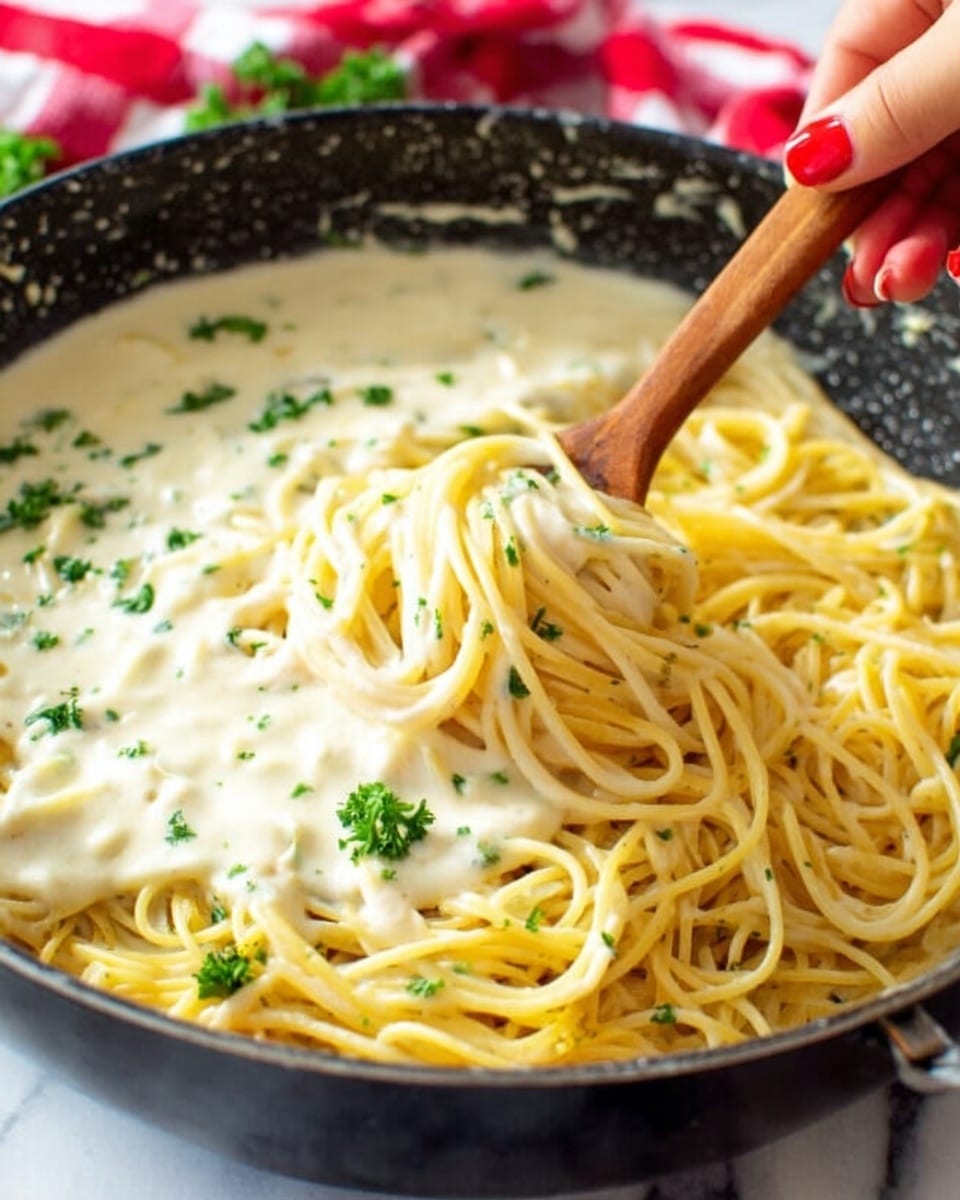 A close-up view of a skillet filled with golden yellow spaghetti pasta, partially covered with a creamy white sauce that looks smooth and thick. Small green parsley leaves are scattered over the sauce and pasta, adding a fresh touch. A woman's hand with red nail polish is holding a wooden spoon, stirring the sauce and pasta together at the edge of the skillet. The skillet's interior is black with some white spots, creating a subtle contrast. The background features a white marbled texture and a red checkered cloth can be partially seen behind the skillet. photo taken with an iphone --ar 4:5 --v 7